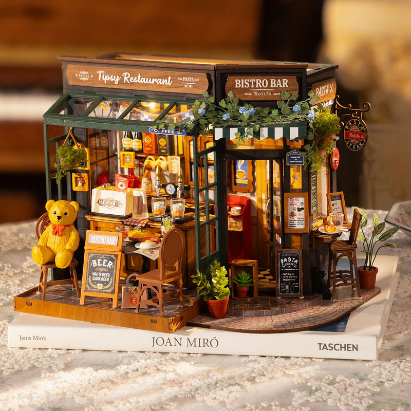 Robotime Tipsy Restaurant, färdigbyggd miniatyrbistro med nalle och krukväxter, uppställd på ett par böcker